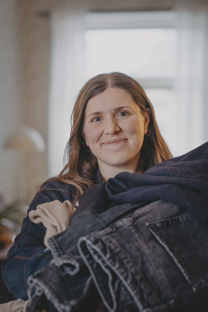 Evelina Albin porträttbild med stenmur, mogna vetefält och lövskog i bakgrunden. Evelina ler och har vinden i håret.